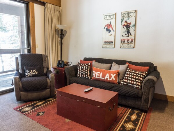 A living room with a leather armchair, a sofa with patterned cushions, a red coffee table, and a lamp. There are two posters on the wall.