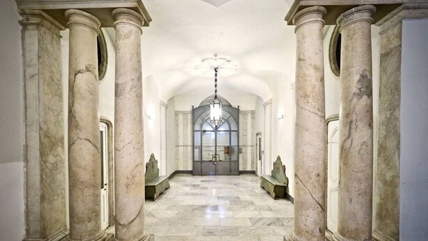 Un vaste hall orné de colonnes de marbre, d’un lustre et d’une grande fenêtre en arc de cercle au fond.