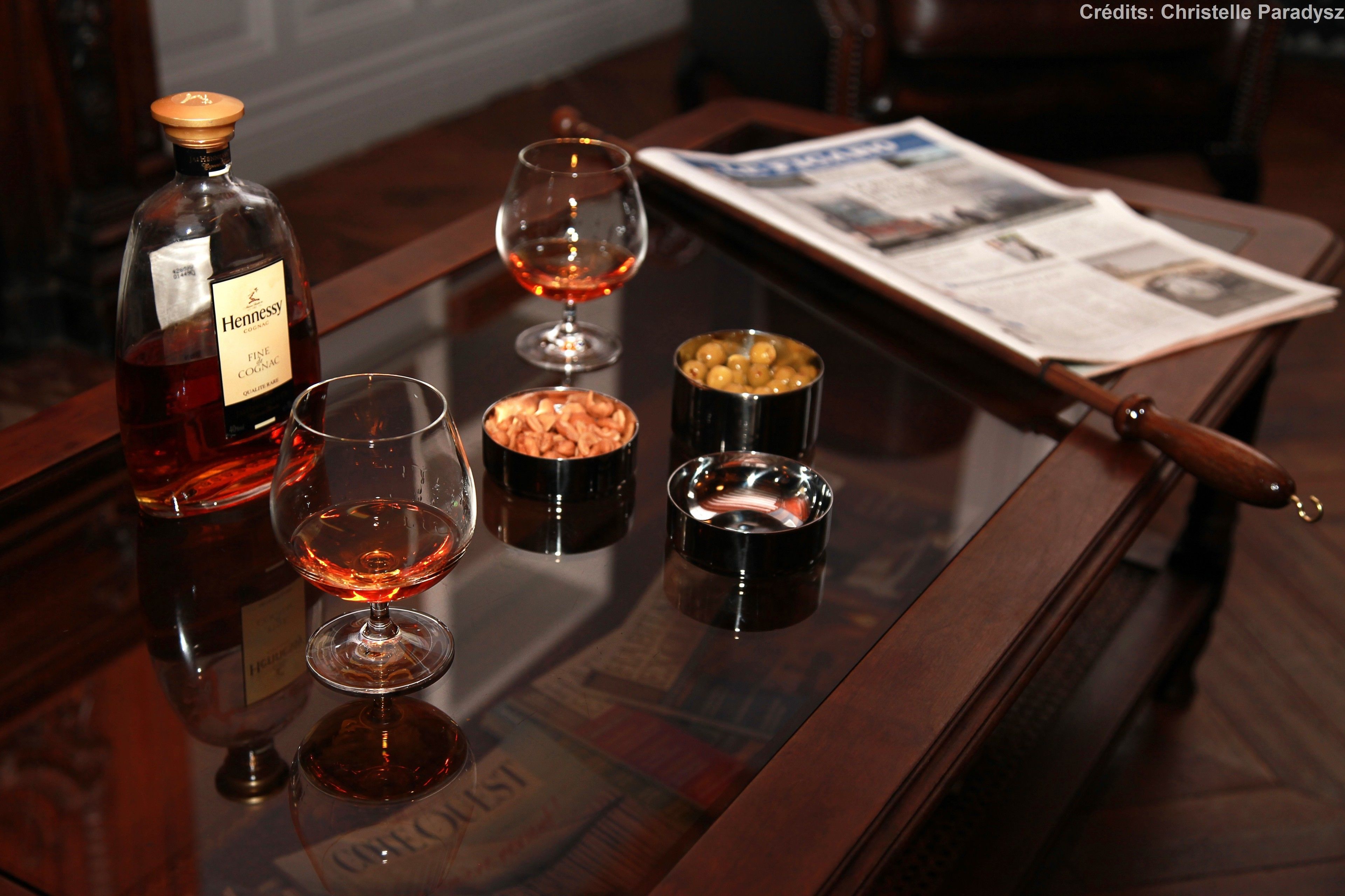 A bottle of Hennessy cognac, two glasses, and small containers of olives and nuts on a wooden table.