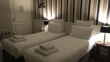Habitación de hotel con dos camas, una puerta, perchero, mesita de noche con lámpara y pared a rayas.