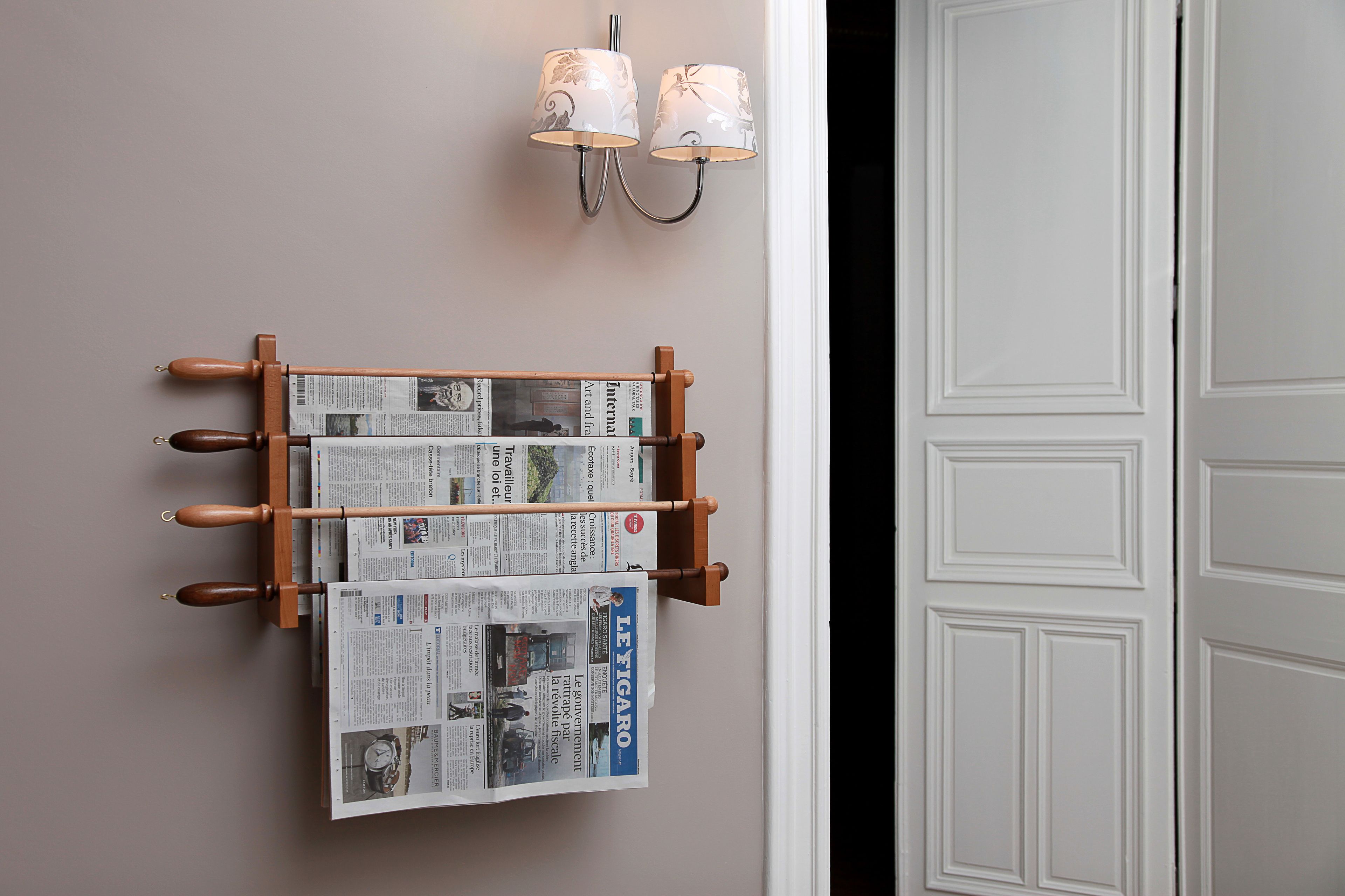 A wall-mounted newspaper rack with several newspapers, a door with paneling, and a wall-mounted light fixture.