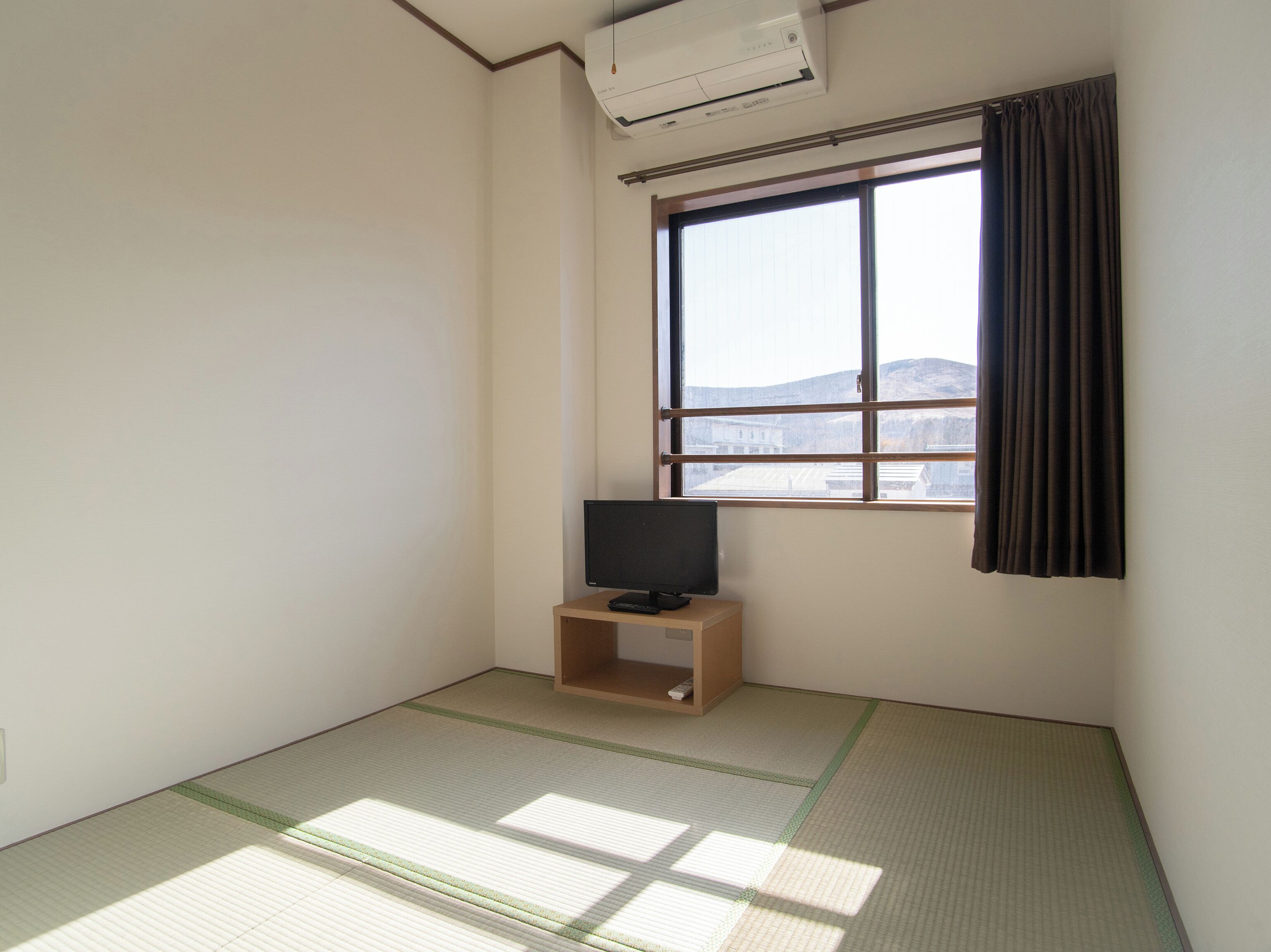 Quarto com piso de tatame, televisão sobre suporte de madeira, janela com vista para um edifício e montanhas, e unidade de ar condicionado no teto.