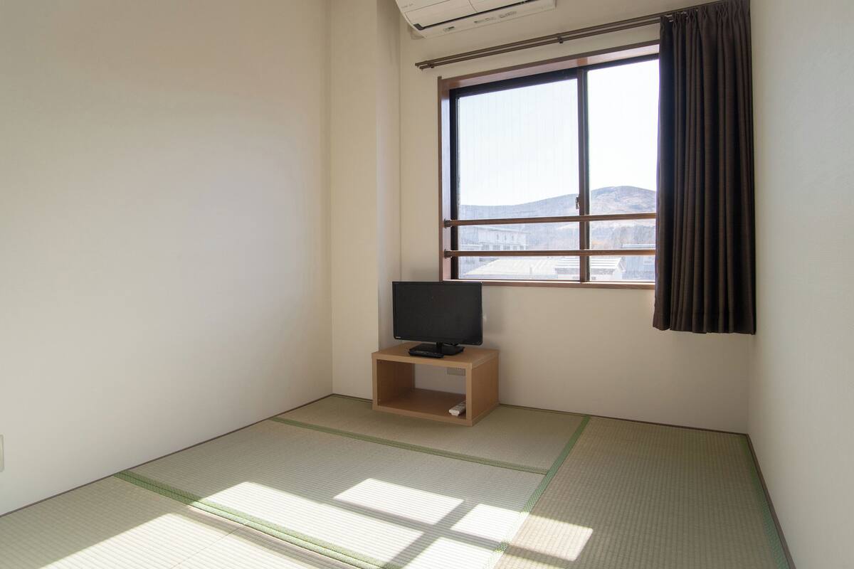 Ein Zimmer mit Tatami-Boden, einem Fernseher auf einem Holzständer und einem Fenster mit Vorhängen.