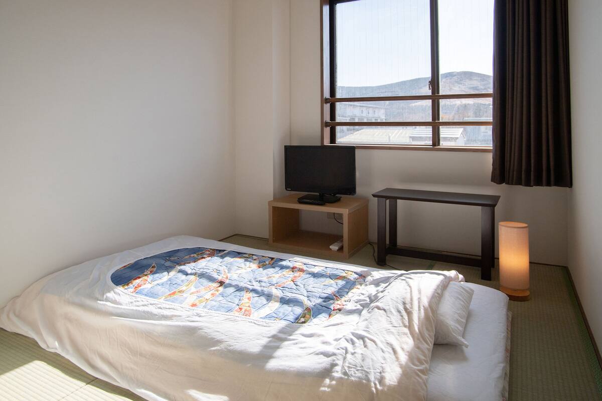 Ein Hotelzimmer mit Bett, Fernseher auf einem Holzgestell, einem kleinen Tisch und einem Fenster mit Vorhängen.