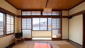 Une pièce de style japonais traditionnel, avec un sol recouvert de tatamis, une table basse en tatami, des portes coulissantes en papier de riz (shoji), un téléviseur posé sur une étagère basse, et une vue sur un lac et des montagnes.
