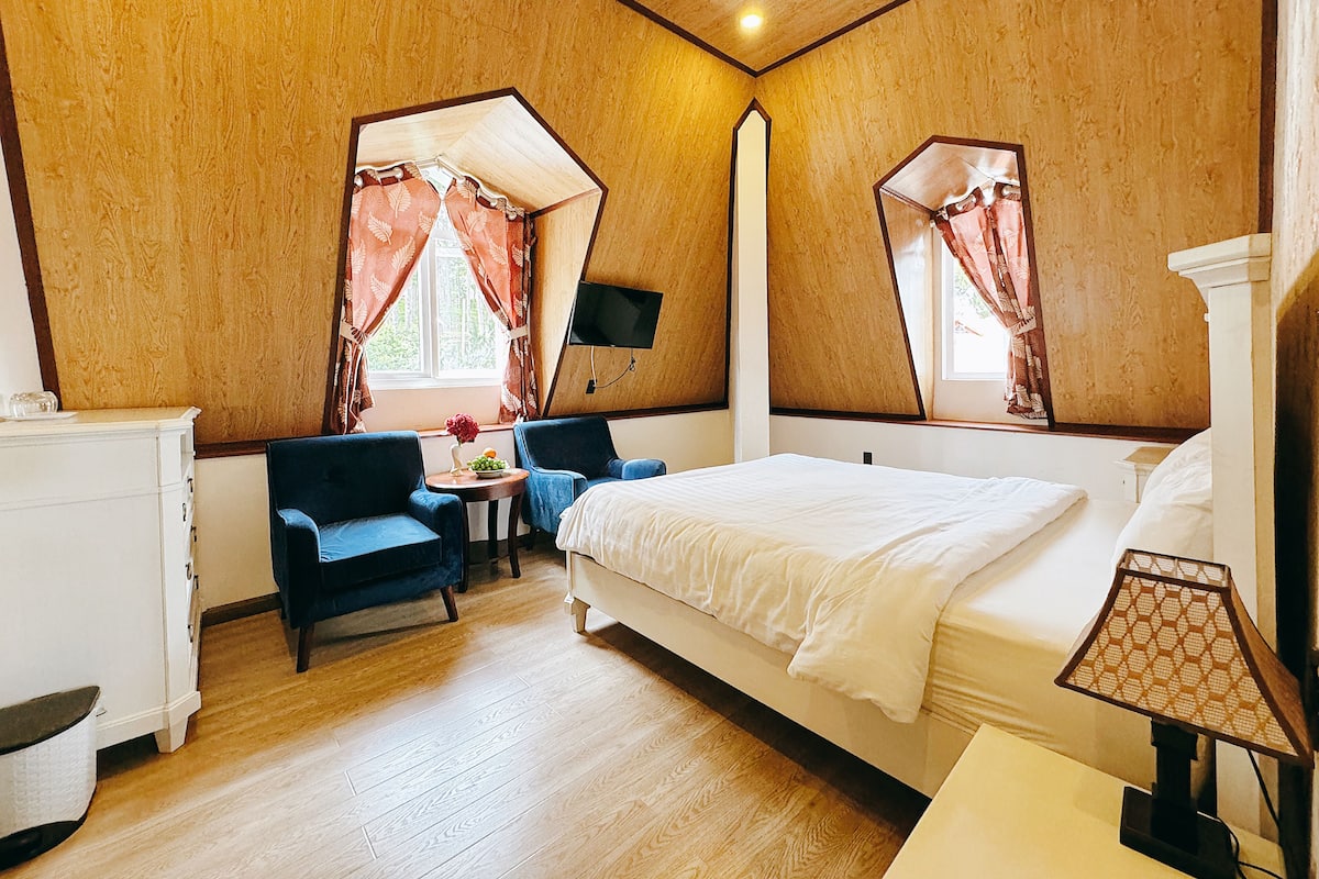 Un dormitorio con cama, dos sillas, una mesita, televisor y lámpara.
