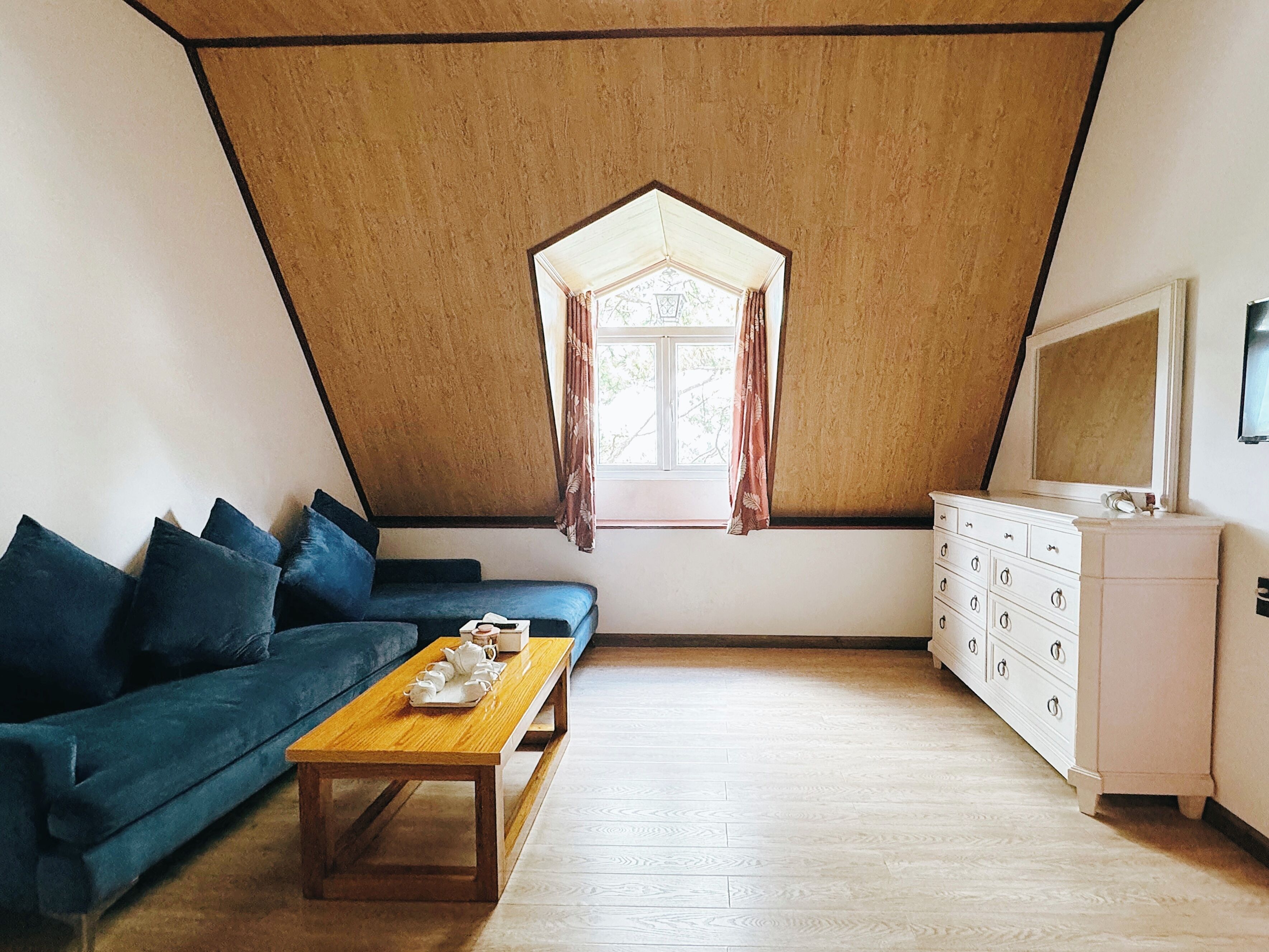 A room with a sloped ceiling, a sofa, a wooden table, a dresser, and a window with curtains.