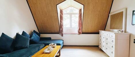 A room with a sloped ceiling, a sofa, a wooden table, a dresser, and a window with curtains.