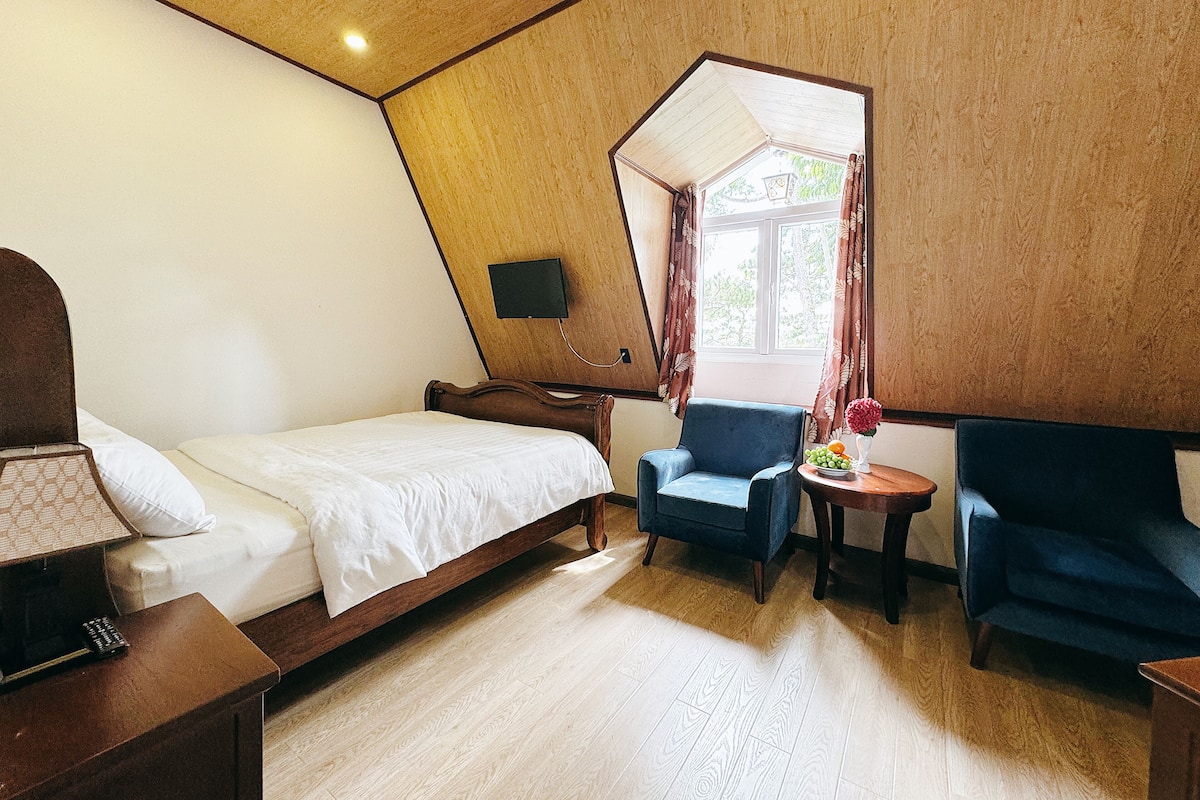 Un dormitorio con cama, una silla, una mesita y televisor montado en la pared.