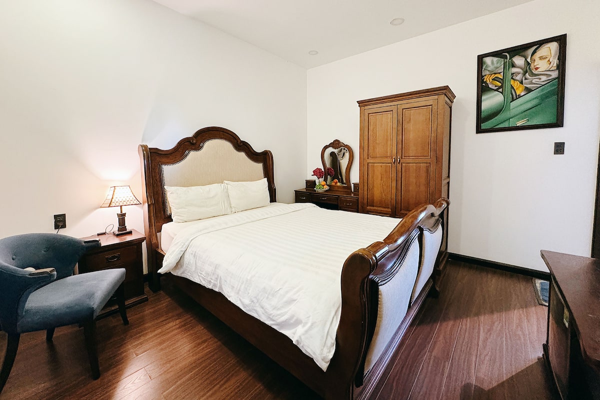 Un dormitorio con una cama de madera, una mesita de noche con lámpara, una silla, un armario y un cuadro en la pared.
