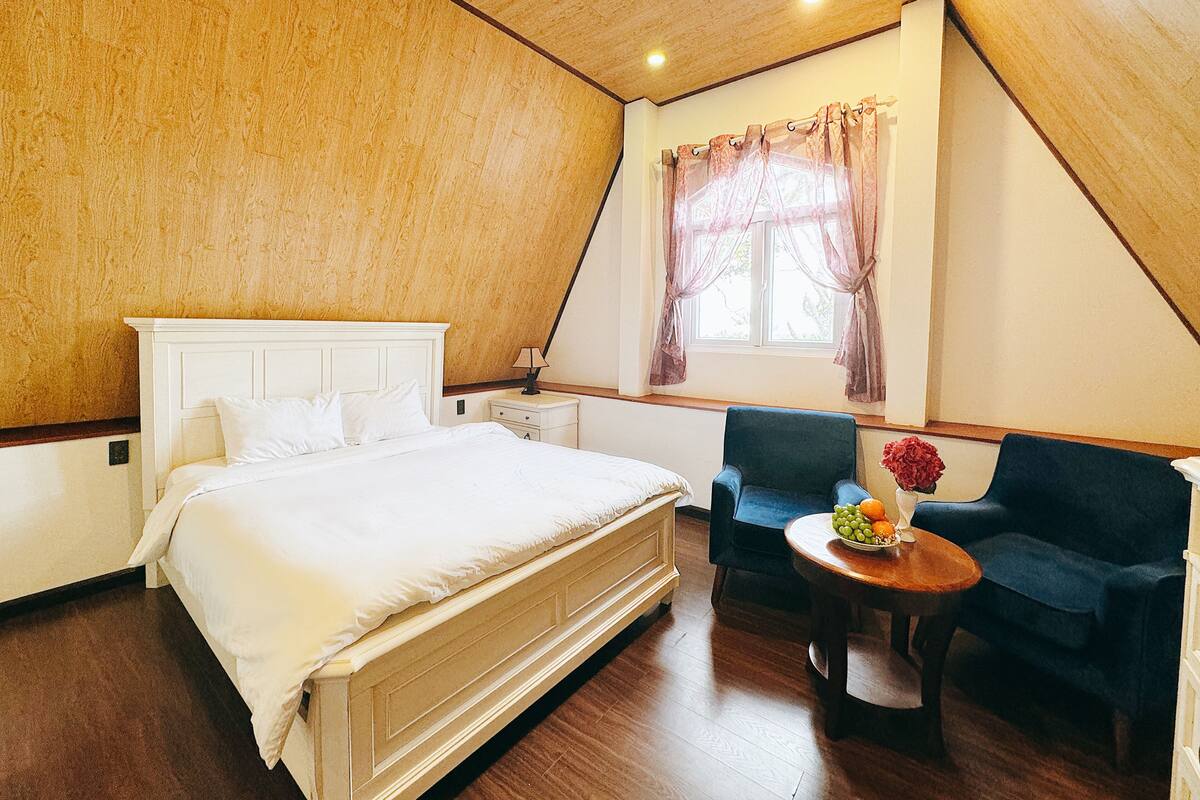 Un dormitorio con una cama grande, una silla, una mesita con fruta y una ventana con cortinas.