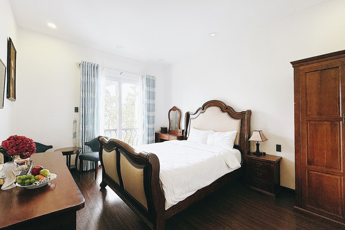 Habitación de hotel con cama de madera, un escritorio con un florero, una silla y una ventana con cortinas.