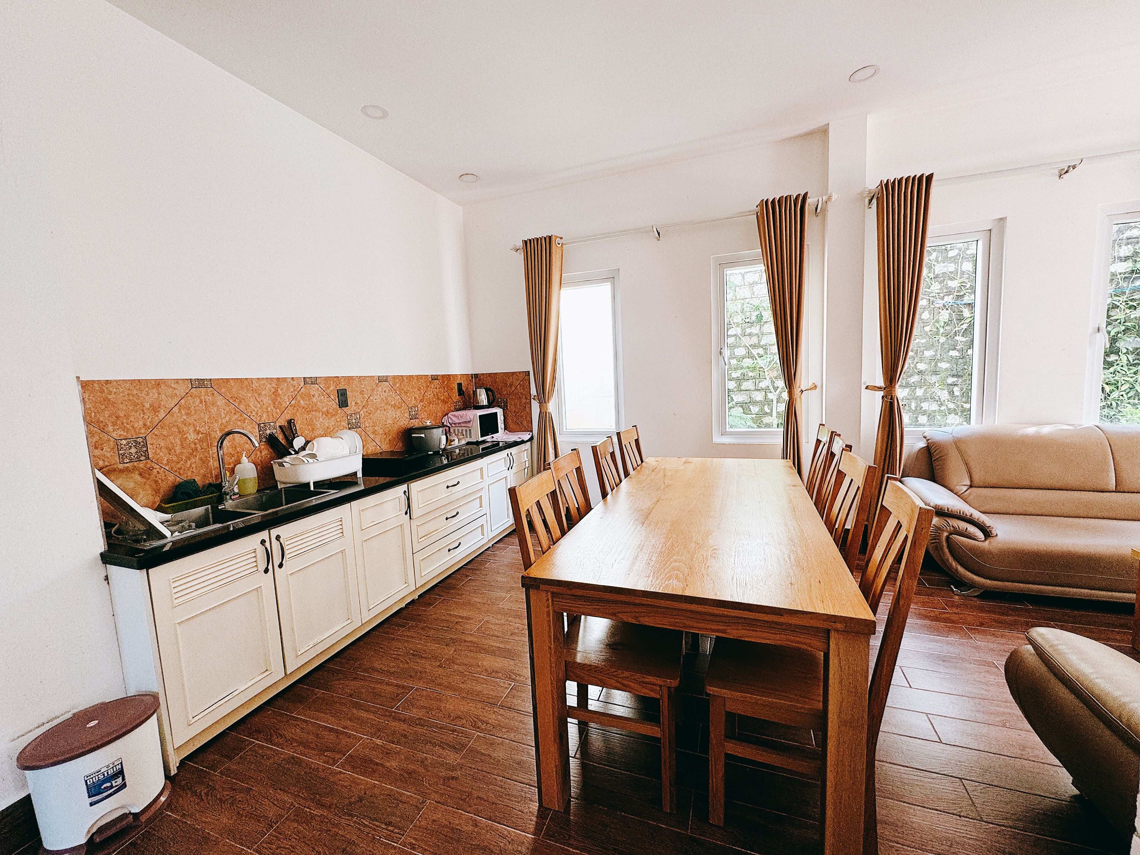 A dining area with a wooden table and chairs, a kitchen with white cabinets, and a window with curtains.