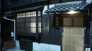 Um edifício tradicional japonês com fachada de madeira escura, telhado telhado e porta de madeira com sistema deslizante. Dois contentores de lixo pretos estão do lado de fora da entrada.