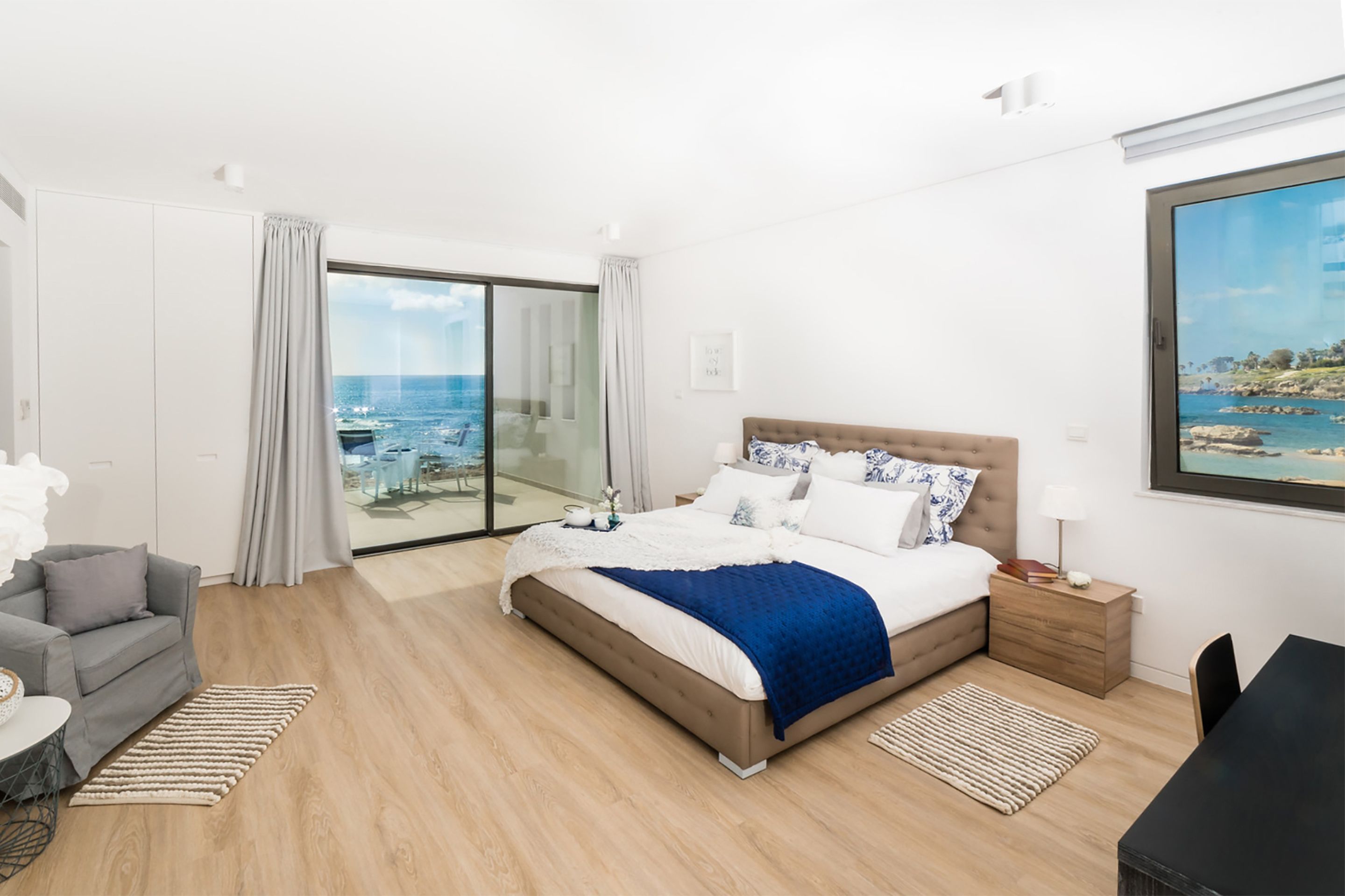 A modern hotel room with a large bed, a grey sofa, a dining table, and a view of the sea through a glass door.