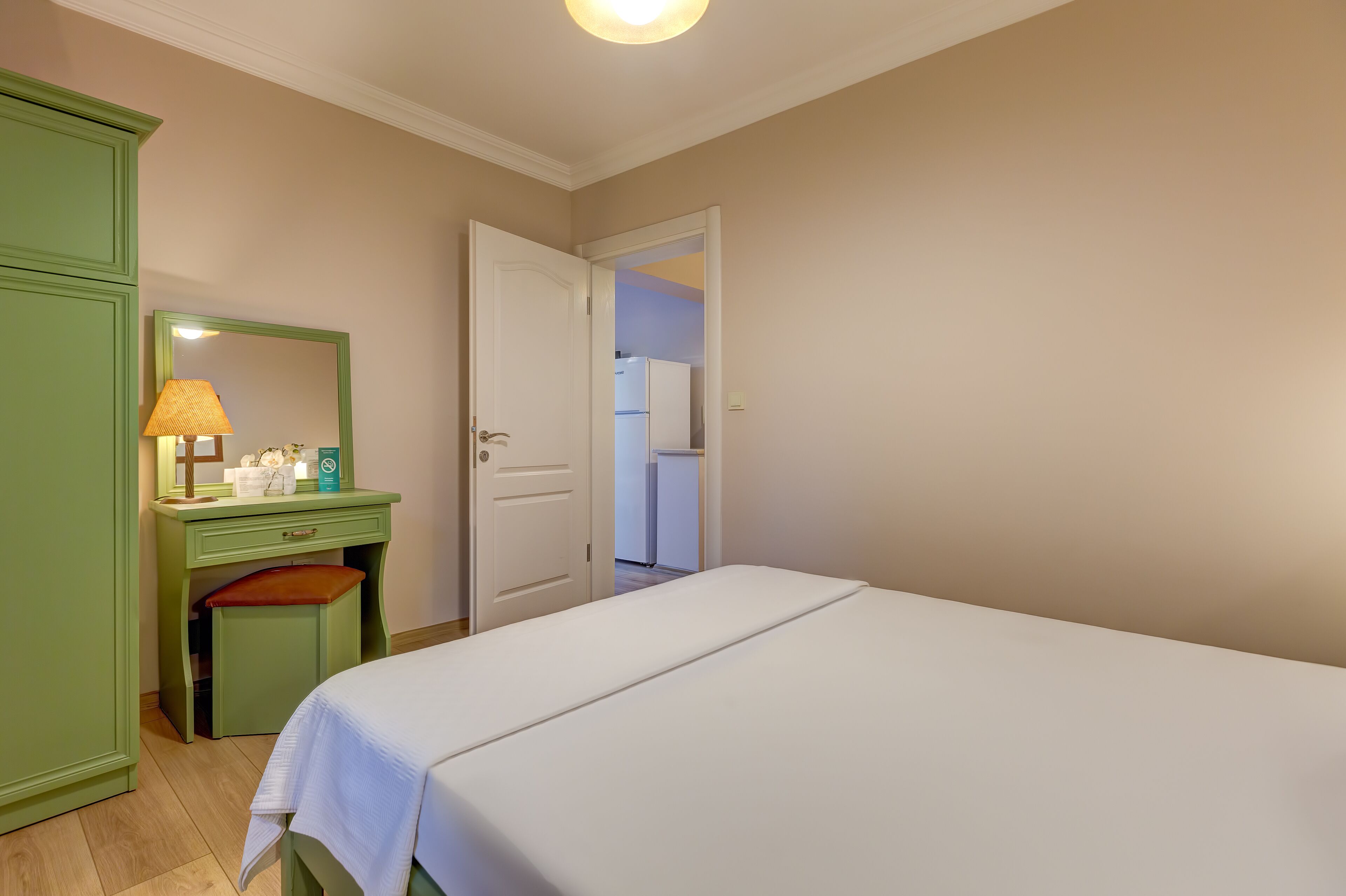 A bedroom with a bed, a green dresser, a mirror, and a door leading to a kitchen.