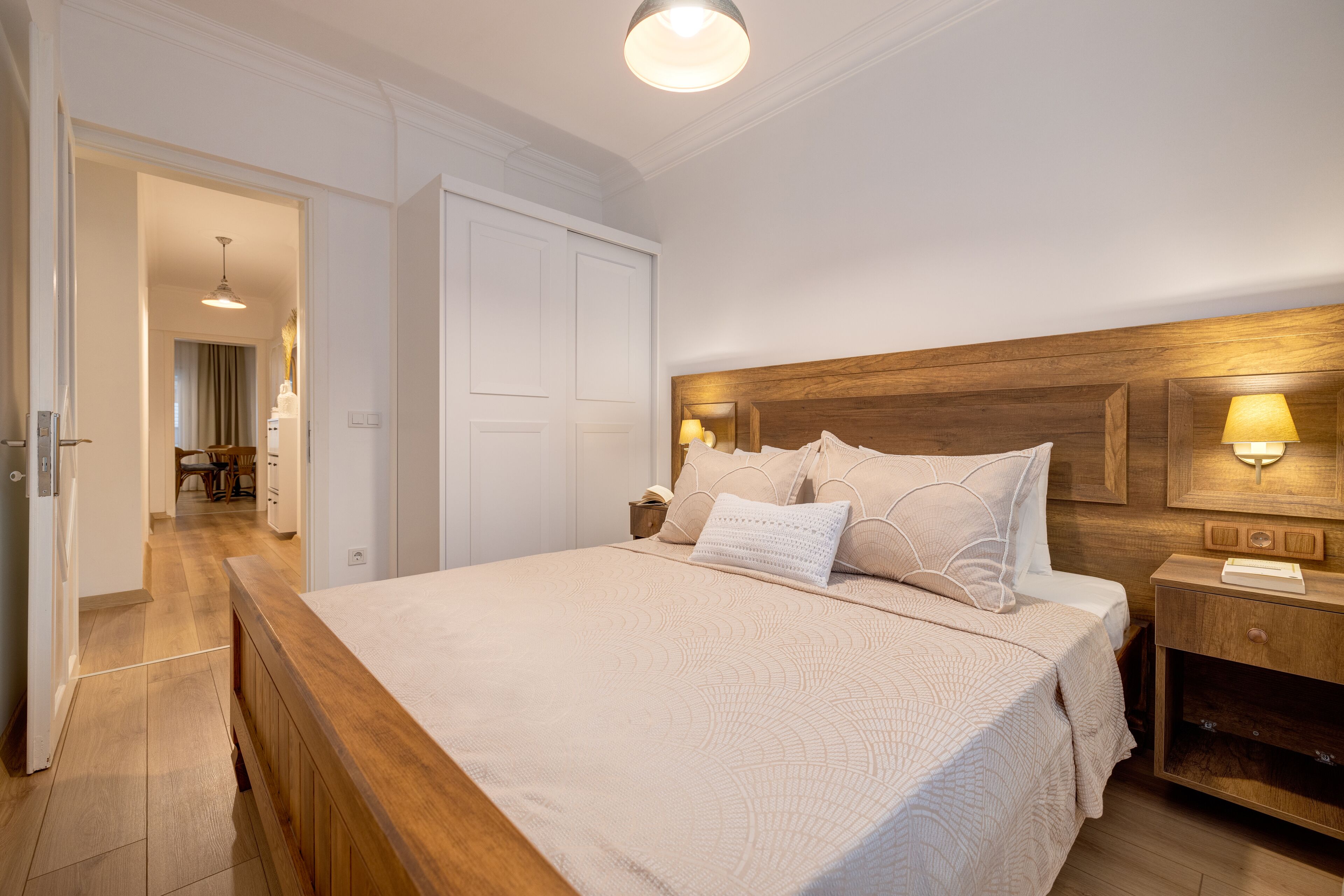 A bedroom with a wooden headboard, a large bed with a beige quilt, two bedside lamps, and a nightstand with a lamp.
