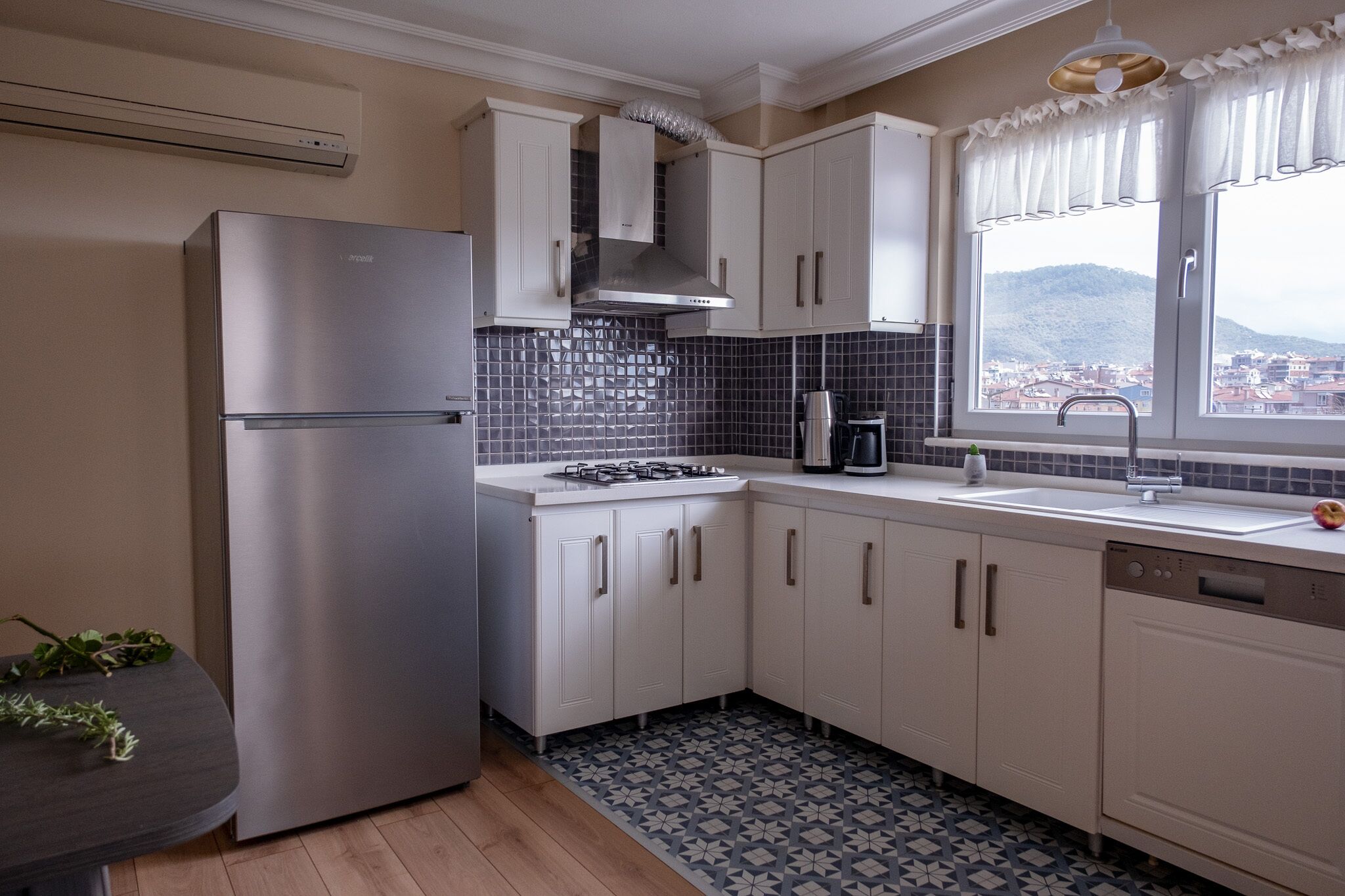 A modern kitchen with white cabinets, a stainless steel refrigerator, a gas stove, and a window with a view of a city and hills.