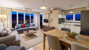 Una sala moderna con sofá, mesa de centro y comedor. Hay una cocina con refrigerador y alacenas. Amplias ventanas ofrecen vistas de la ciudad al atardecer.
