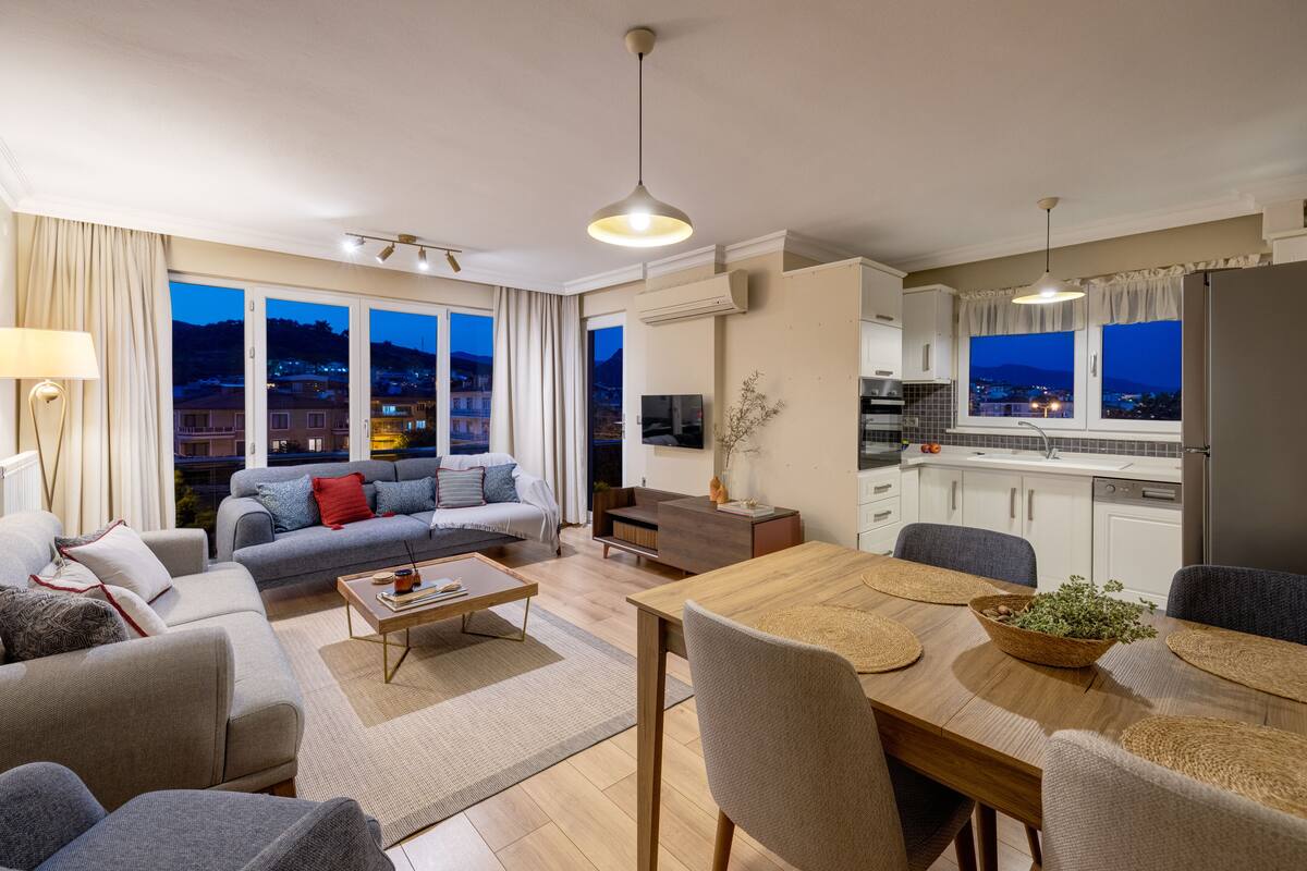 Una sala moderna con sofá, mesa de centro y comedor. Hay una cocina con refrigerador y alacenas. Amplias ventanas ofrecen vistas de la ciudad al atardecer.
