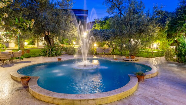 Una piscina circular con una fuente central, rodeada de árboles y plantas en macetas, con mobiliario exterior y un edificio al fondo.