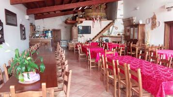Une salle à manger avec de longues tables et des chaises en bois, recouvertes de nappes rouges. Un téléviseur est fixé au mur et un escalier mène à un niveau supérieur.