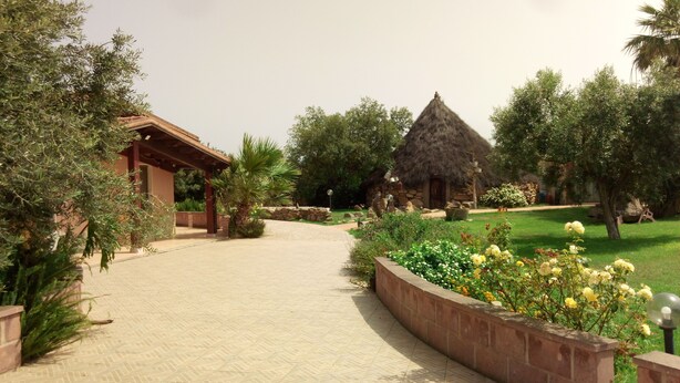 A pathway leading to a rustic building with a thatched roof, surrounded by lush greenery and flowering plants. - Agriturismo Is Scalas (Assemini)