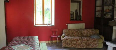 A room with a red wall, a checkered floor, a floral sofa, a wooden table, and a window with a view of greenery.