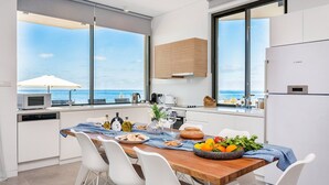 A modern kitchen with a view of the sea, featuring a wooden table set with food, white chairs, and a large window. - Villa Lara (Kouklia)