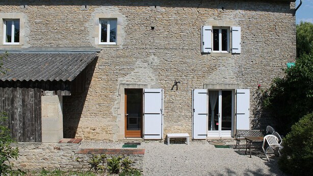 A stone building with two windows, a wooden door, and a small outdoor seating area. - Gîte- La Ferme des Chataigniers (Vienne-en-Bessin)