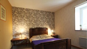 A bedroom with a bed, two bedside tables, a window with a plant, and a floral wallpaper. - Gîte- La Ferme des Chataigniers (Vienne-en-Bessin)