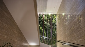 A modern interior with a glass staircase, a central garden area with plants and wooden elements, and a view of greenery through a large window.