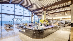 A spacious hotel lobby with a large buffet area, high ceilings, and large windows offering a view of the ocean.