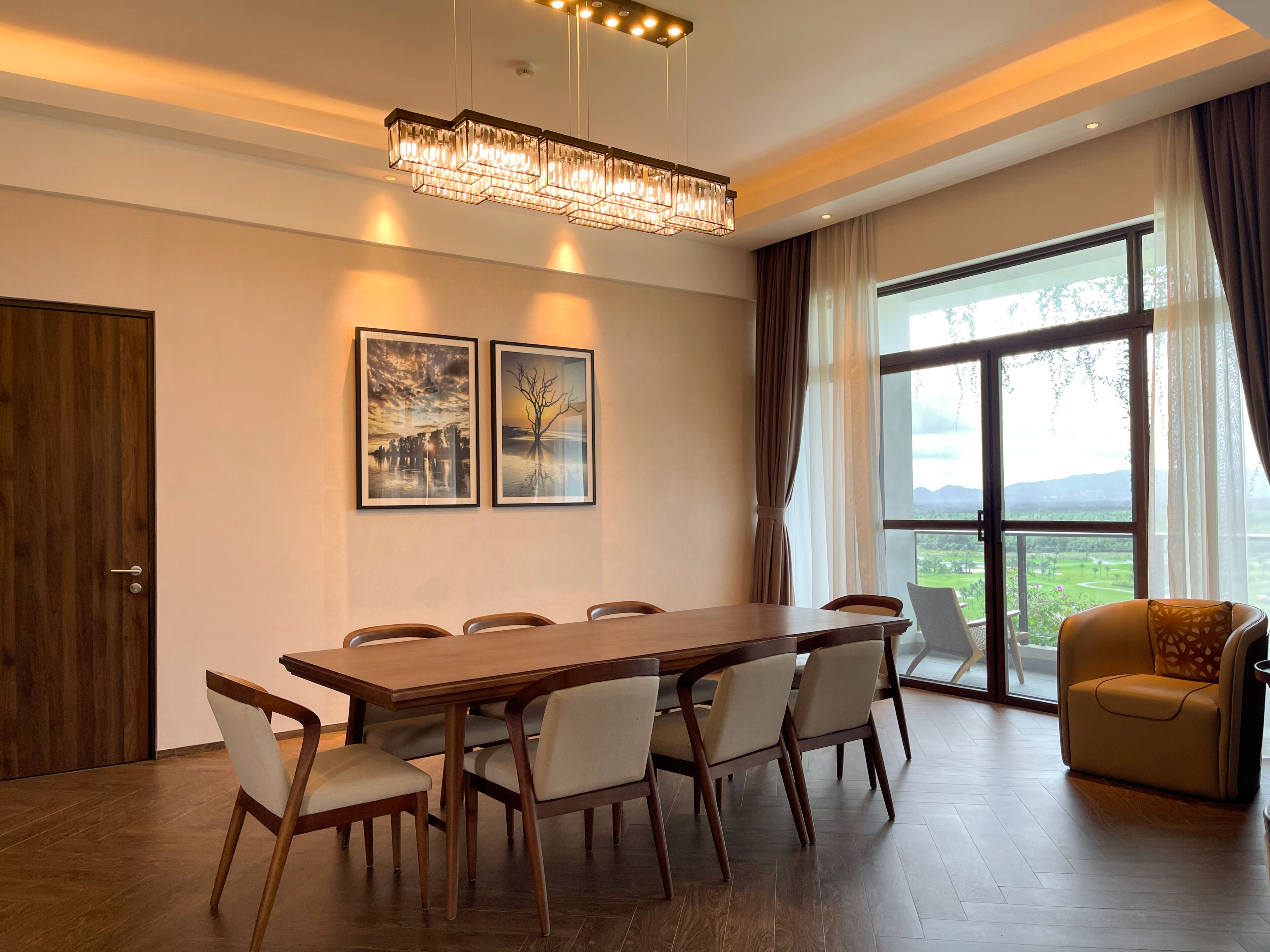 Un espace de repas moderne avec une table et des chaises en bois, un lustre et de grandes fenêtres donnant sur l’extérieur.