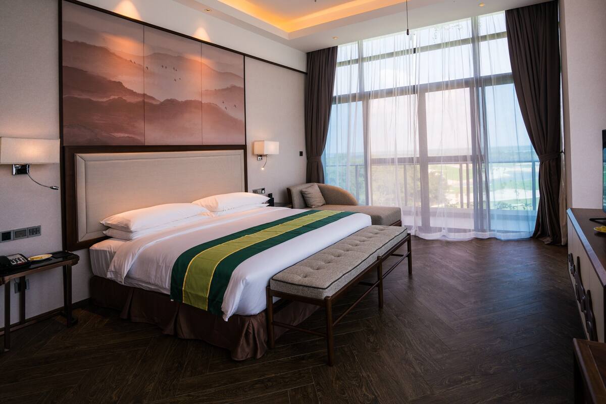 Una habitación de hotel con una cama grande, un banco, un escritorio con televisor, una ventana con cortinas y un cuadro en la pared.