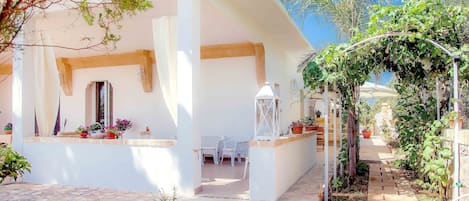 A white building with a covered patio area, outdoor seating, and a garden path.