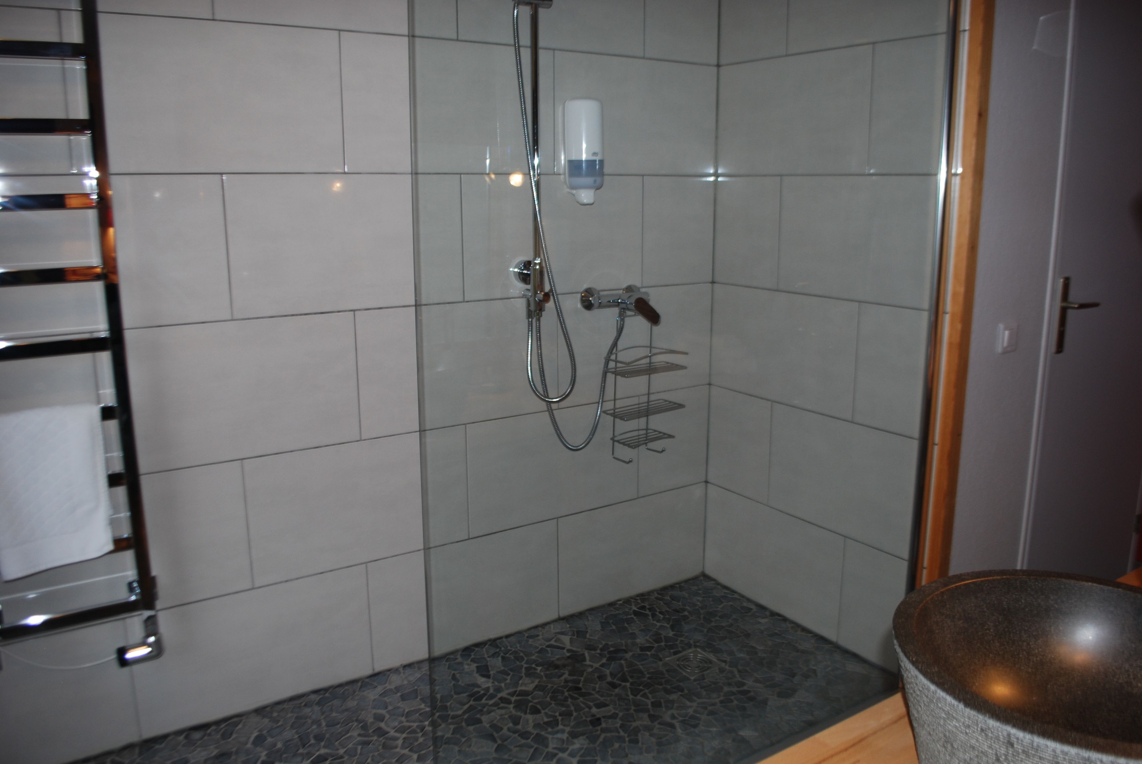A shower with a handheld showerhead, a wall-mounted soap dispenser, and a tiled floor.