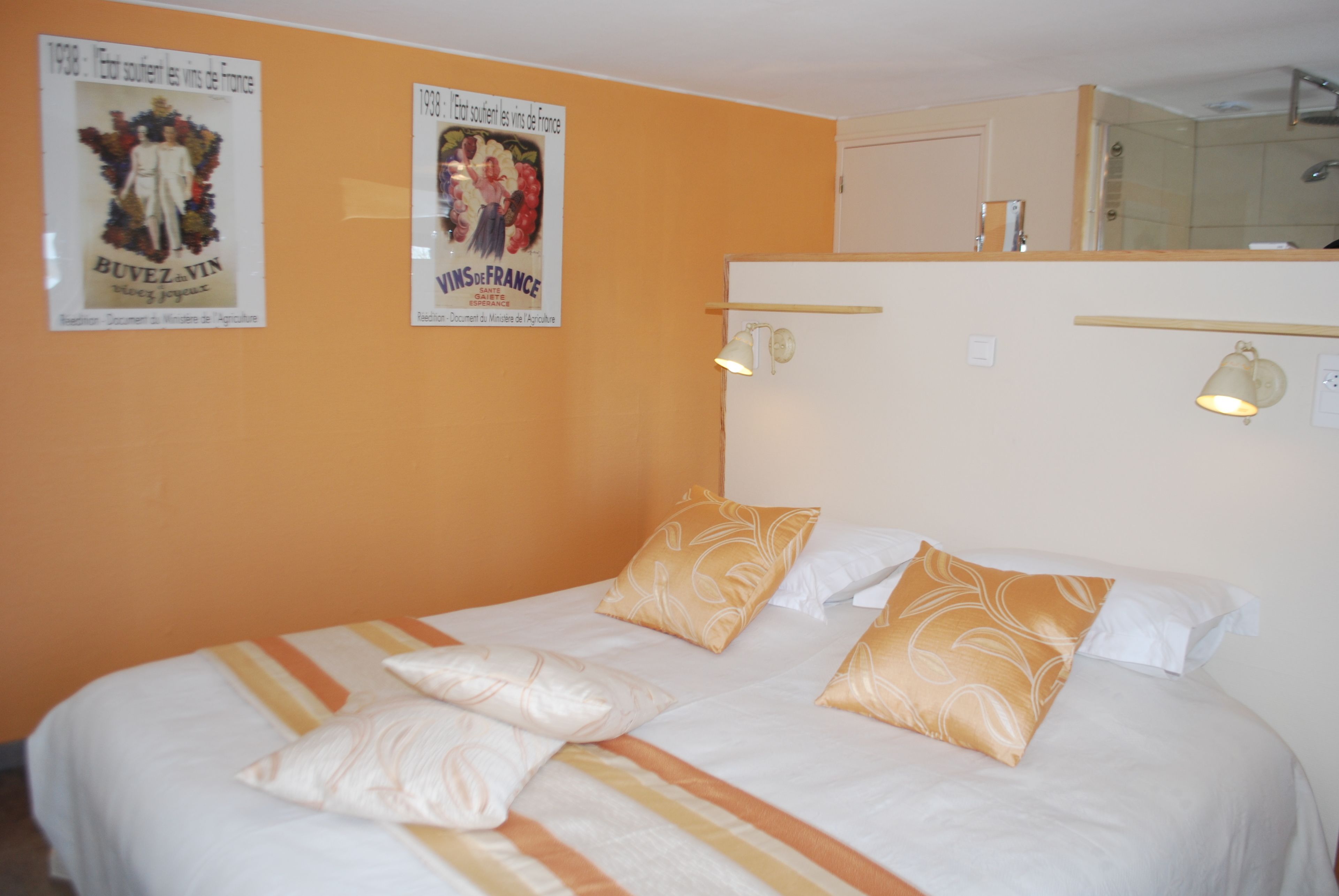 Habitación de hotel con una cama, dos carteles en la pared, un espejo sobre la cama y una pequeña estantería con una lámpara.