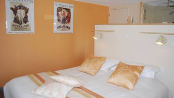 Habitación de hotel con una cama, dos carteles en la pared, un espejo sobre la cama y una pequeña estantería con una lámpara.
