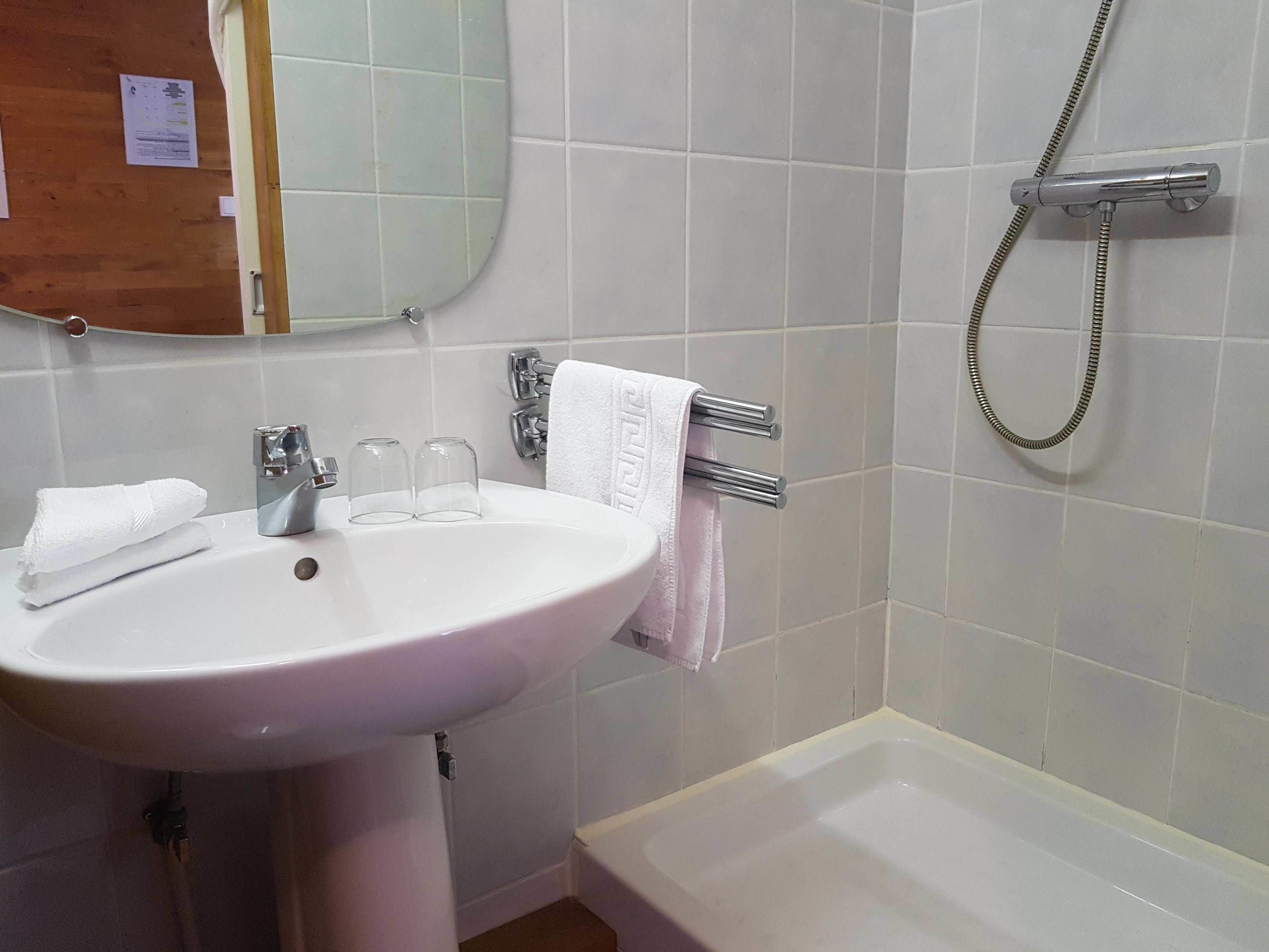 Un baño con un lavamanos blanco, dos vasos de vidrio transparente, una toalla, un espejo y una ducha.