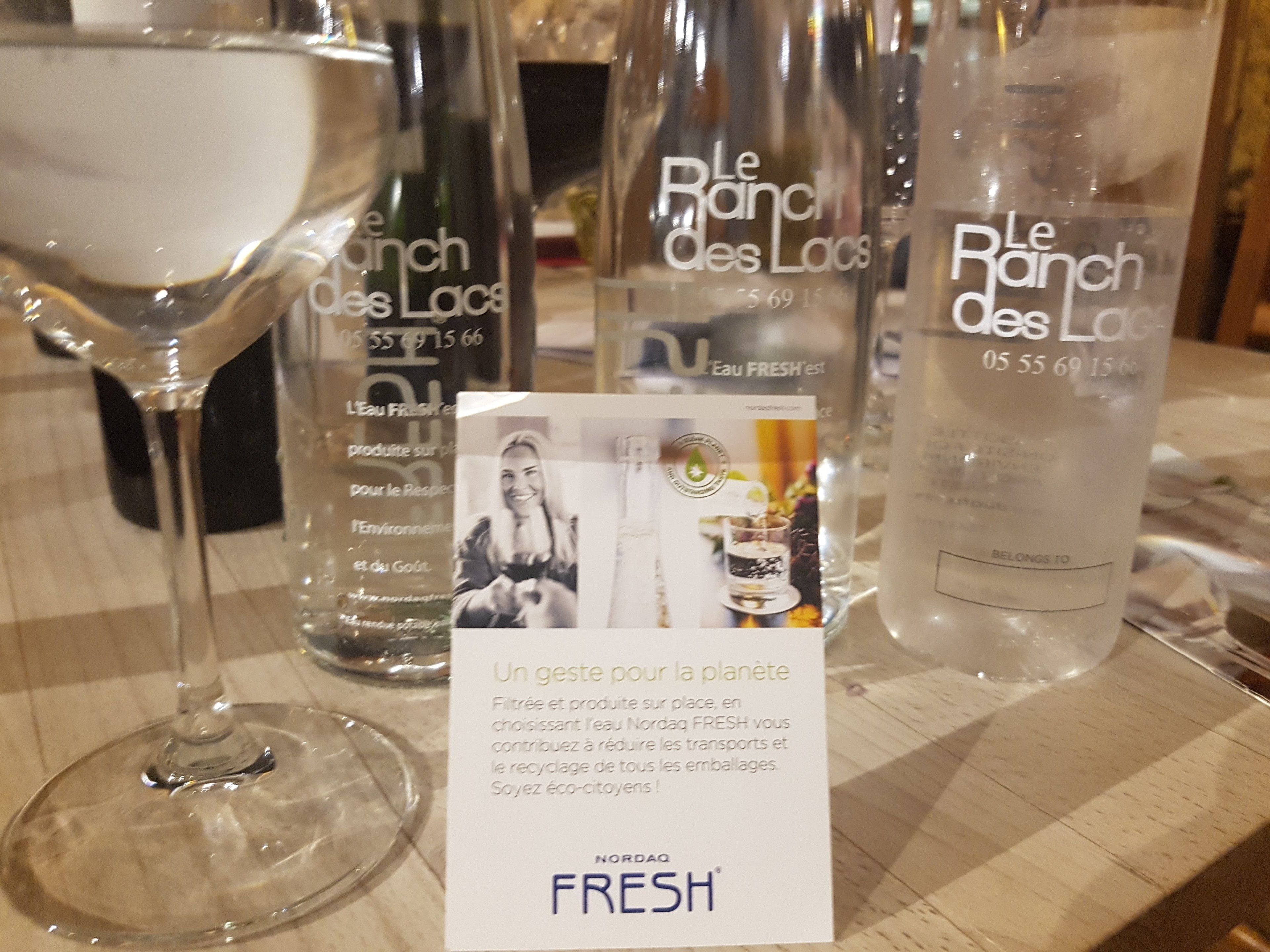Uma mesa com uma taça de martini, três garrafas de água Le Ranch des Lacs, e um cartão promovendo práticas ecológicas.