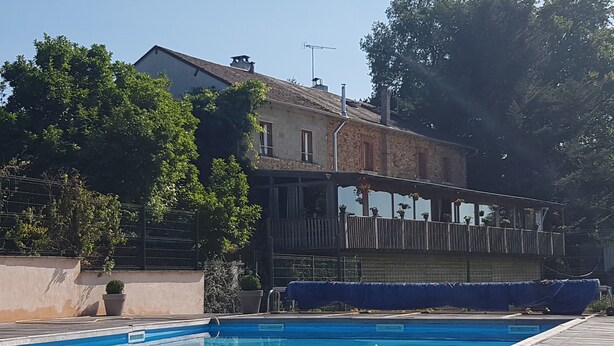 A swimming pool with a wooden deck, a stone building with a balcony, and a fence with greenery. - Le Ranch des Lacs, Hôtel Restaurant à Augne (Augne)