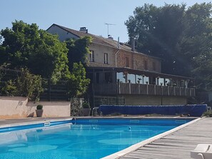 Una piscina amb una terrassa de fusta, un edifici de pedra amb un balcó, i una tanca amb vegetació.