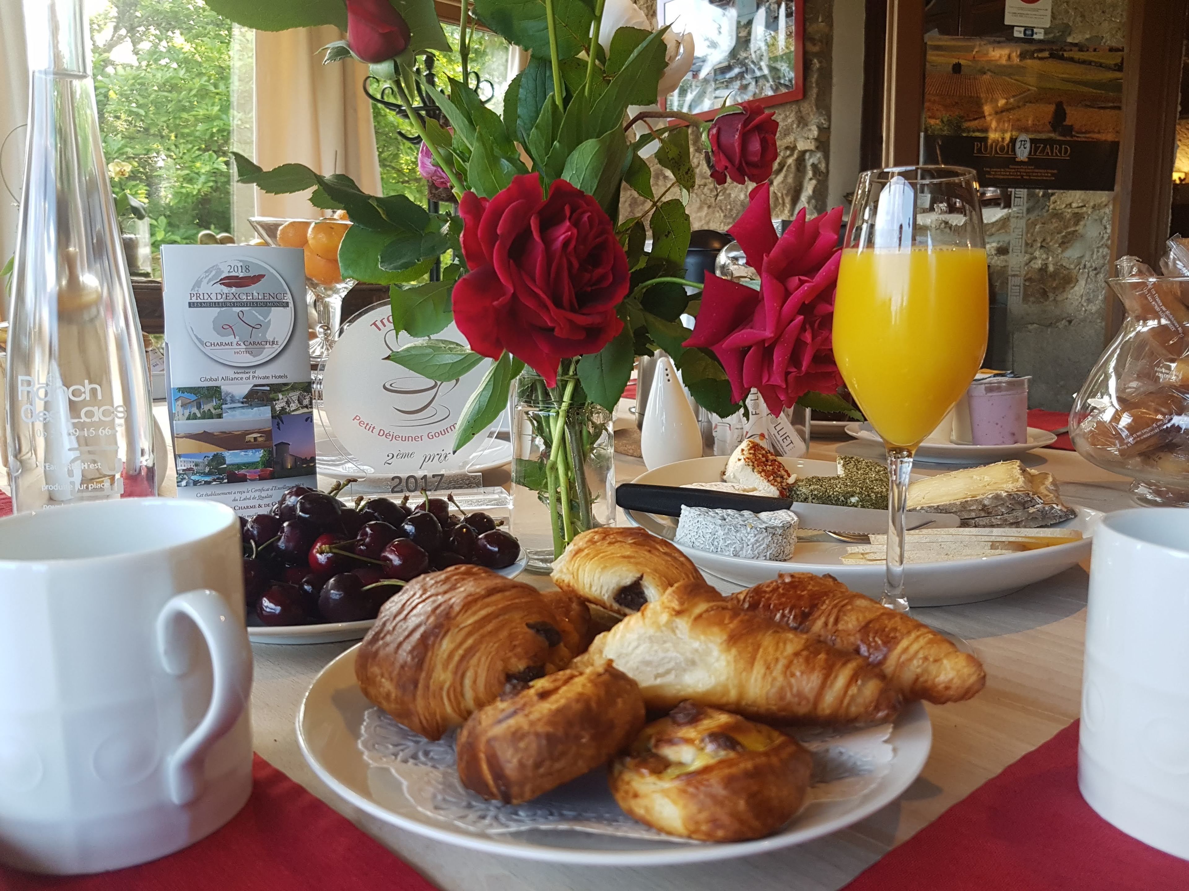 Una mesa de desayuno con croissants, cerezas, un vaso de jugo de naranja y un florero con rosas rojas.