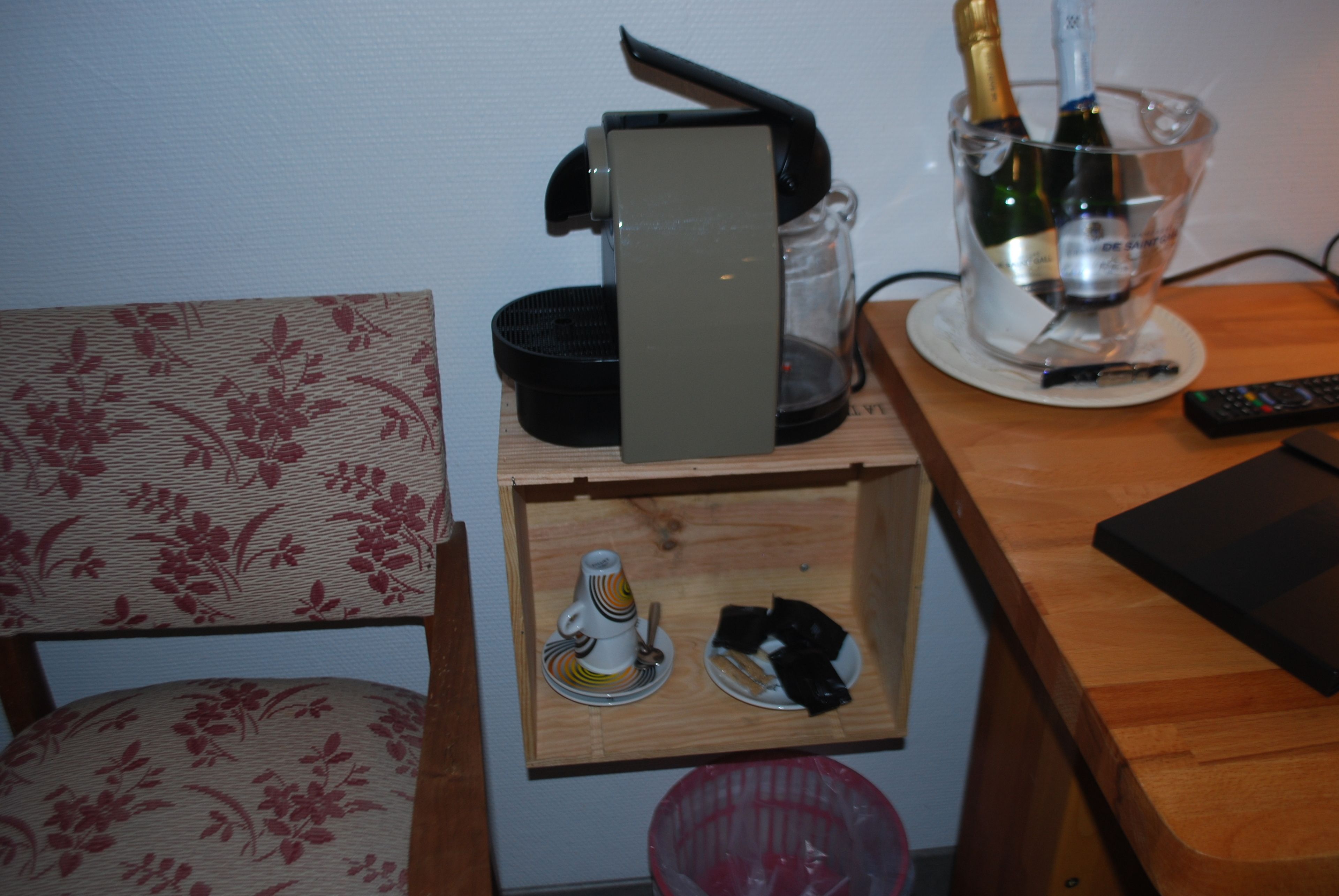 A wooden side table with a remote control, a glass of wine, and a small drawer containing a cup and saucer.