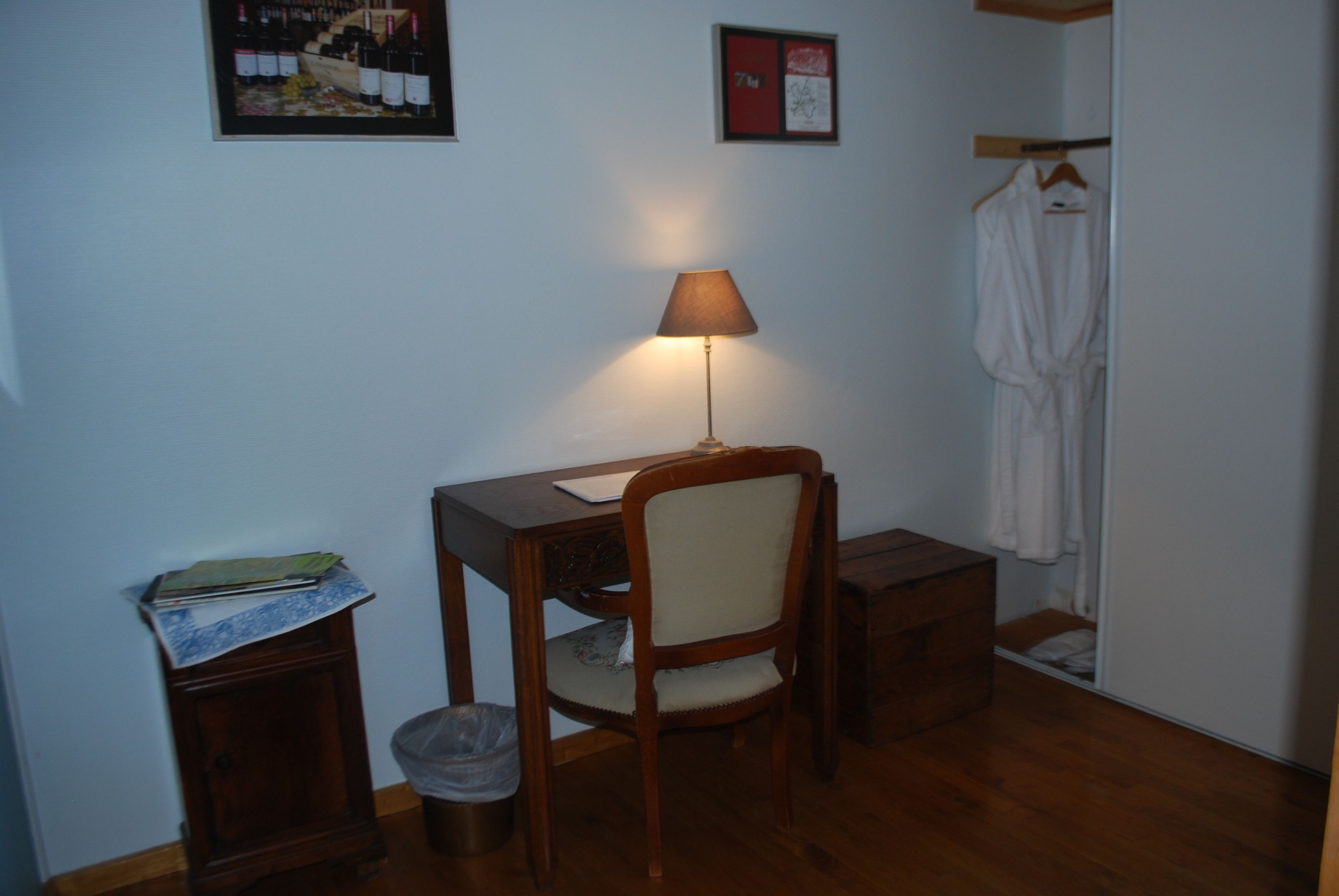 Una stanza piccola con una scrivania in legno, una sedia, una lampada, un accappatoio appeso a un appendiabiti, un cestino e un quadro incorniciato al muro.