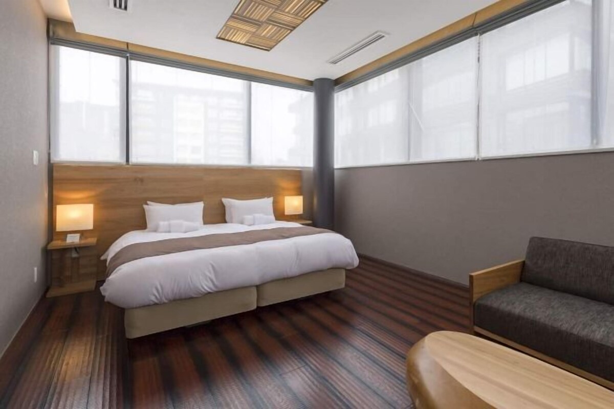 A modern hotel room with a large bed, a sofa, and a view of the city through large windows.