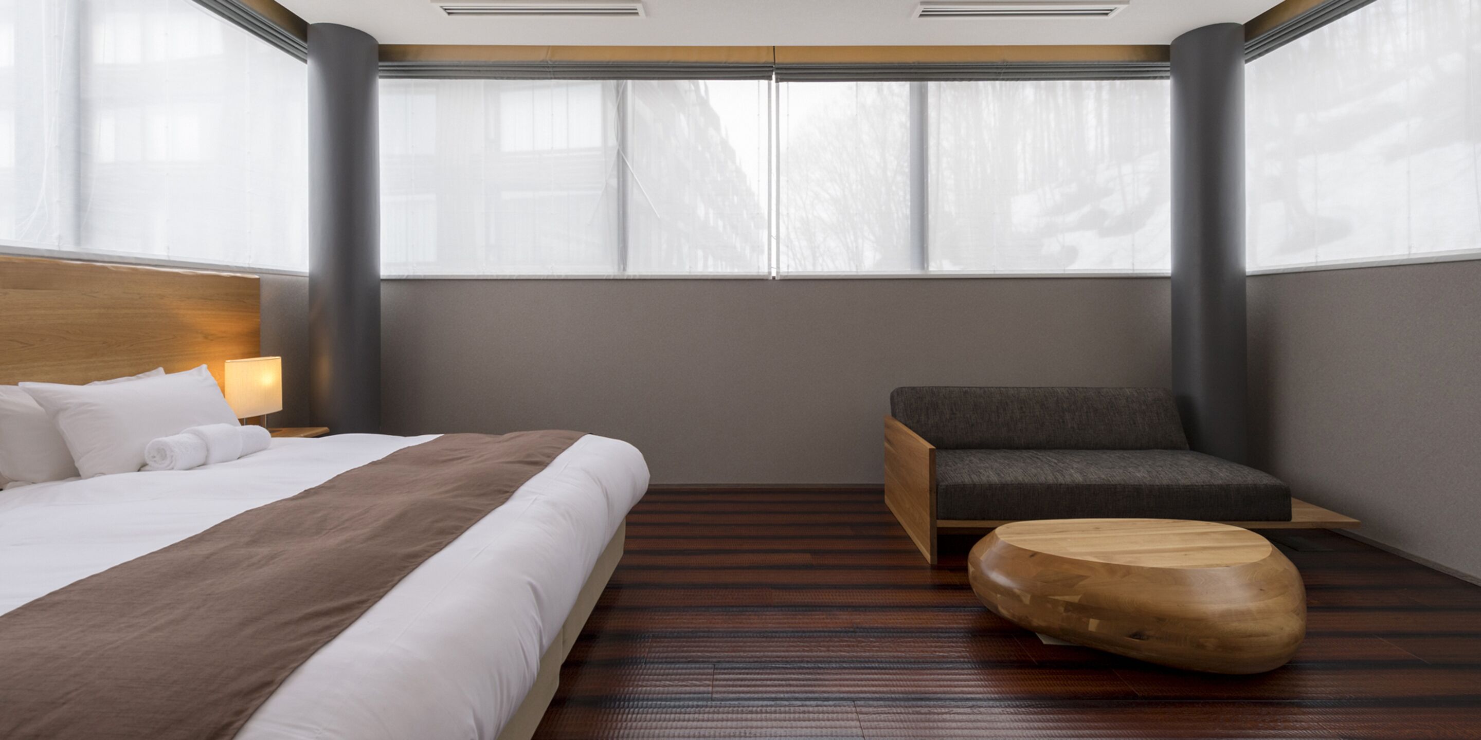 Une chambre d’hôtel moderne avec un grand lit, une table de chevet en bois, un fauteuil gris et un pouf en bois.