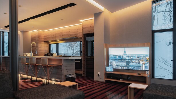 Una sala de estar moderna con un televisor grande, asientos cómodos, una zona de cocina y ventanas amplias con vistas a la ciudad.