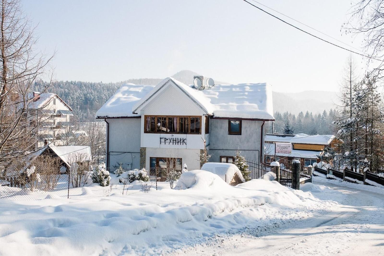 A two-story building with a sign reading "ГРУНИК" in Cyrillic script, surrounded by snow-covered ground and trees.